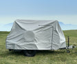 A small trailer with a Hero Trailer Cover by Paha Que Custom, made from heavy-duty MagnaTuff fabric, is parked on green grass under a hazy blue sky, with mountains faintly visible in the background.