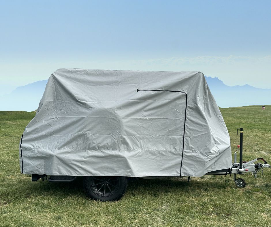 A small trailer with a Hero Trailer Cover by Paha Que Custom, made from heavy-duty MagnaTuff fabric, is parked on green grass under a hazy blue sky, with mountains faintly visible in the background.