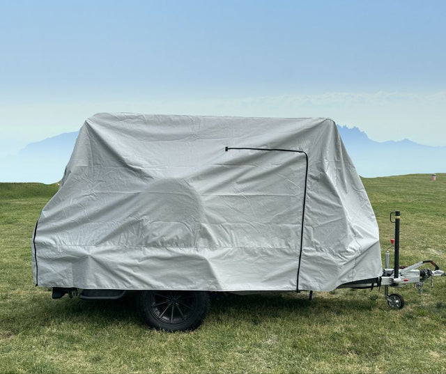 A small trailer with a Hero Trailer Cover by Paha Que Custom, made from heavy-duty MagnaTuff fabric, is parked on green grass under a hazy blue sky, with mountains faintly visible in the background.