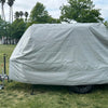 A Squaredrop Trailer covered with the Paha Que Custom 6 x 13 Heavy Duty MagnaTuff Fabric trailer cover stands on grass in a park, with trees and palm trees visible under a partly cloudy sky.