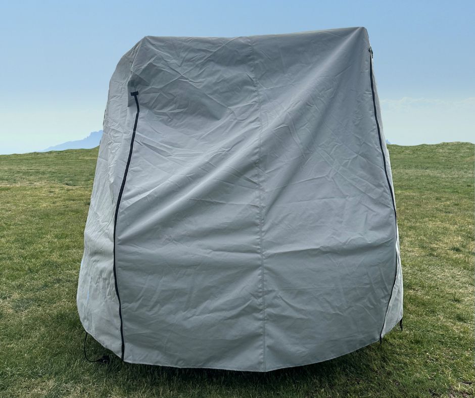 The Hero Trailer Cover by Paha Que Custom, made with heavy duty MagnaTuff fabric for weather-proof protection, is zipped and creased as it stands on a grassy field beneath a clear blue sky with faint mountains in the background.