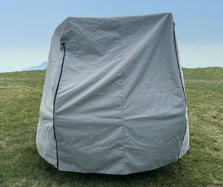 The Hero Trailer Cover by Paha Que Custom, made with heavy duty MagnaTuff fabric for weather-proof protection, is zipped and creased as it stands on a grassy field beneath a clear blue sky with faint mountains in the background.
