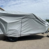 A Hiker Trailer is parked on pavement near industrial buildings, protected by a Paha Que Custom Hiker Trailer Covers Custom Fit Outdoor Protection. Only the trailer’s hitch and one wheel are visible under a partly cloudy sky.