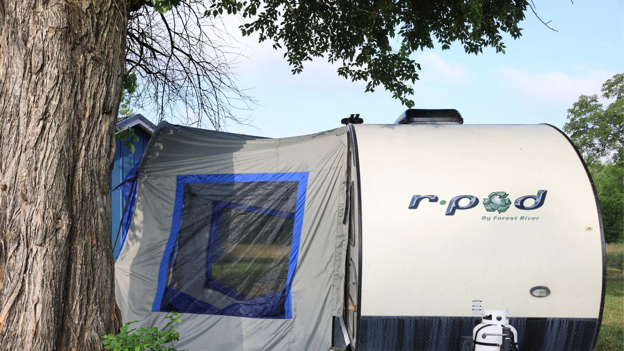 A Forest River R-Pod Trailer Side Tent by PahaQue for Enhanced Camping Comfort is pitched under a large tree, showcasing its blue-trimmed window and easy setup with camping gear—perfect for outdoor adventures. Brand: Paha Que Custom.
