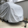 A compact camper with a Paha Que Custom NuCamp TAB 360 CS Trailer Cover by PahaQue, designed for heavy-duty, weatherproof protection, is parked on a concrete driveway by a metal building, with wheel chocks securing its tires.