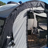 A silver Little Guy Mini Max Side Tent with black trim by PahaQue Wilderness is attached to a camper. Mesh windows and an open door reveal a folding chair inside, set up near a road on a sunny day.
