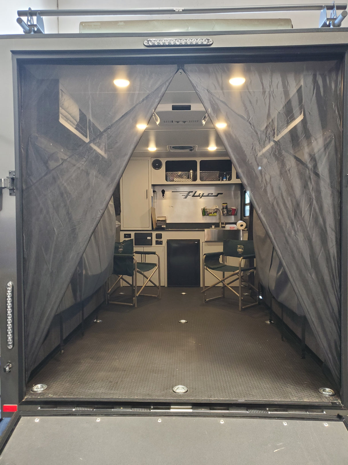 Interior of a camper trailer with Paha Que Custom Mesh Door/Solid Room Divider open for insect protection, featuring two green folding chairs, a kitchenette with cabinets, sink, stove, and overhead lighting along white walls.