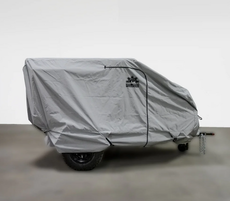 A small off-road trailer is fully protected by the Paha Que Custom Off Grid Trailer Covers Premium Protection for Most OffGrid Trailers, a fitted gray, weather-resistant cover with logo and zipper, parked indoors with hitch and chains partly visible.