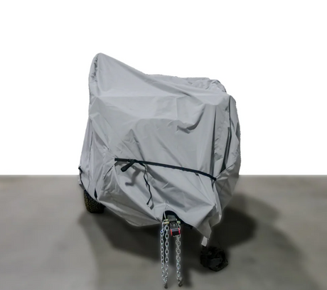 A Paha Que Custom Off Grid Trailer Covers Premium Protection for Most OffGrid Trailers sits on a concrete floor with tow hitch and safety chains visible, showcased against a plain white background for reliable trailer protection.