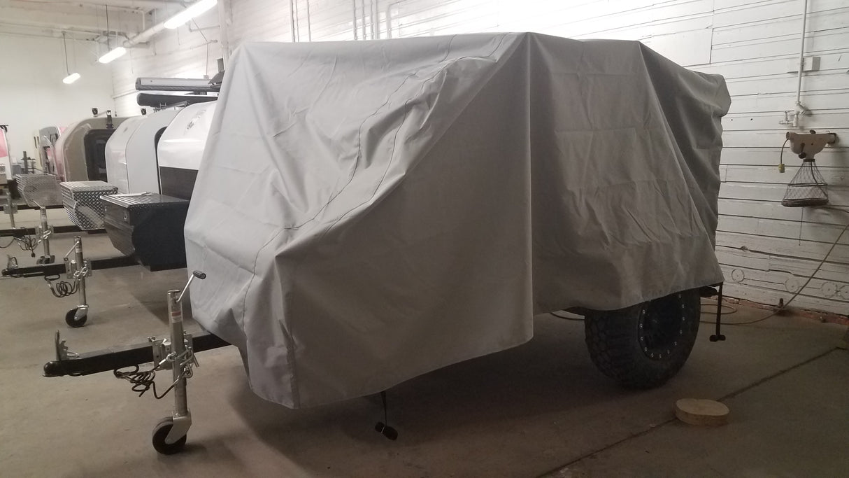 A trailer covered with a PahaQue Wilderness Sherpa Trailer Cover, made from durable MagnaTuff fabric, is parked indoors near a white wall, while two uncovered trailers can be seen in the background.