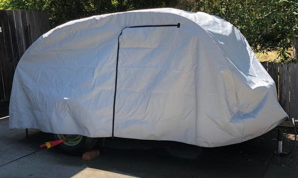A So-Cal Teardrops Sierra Trailer is protected by a fitted gray So-Cal Teardrops Sierra Trailer Cover from PahaQue Wilderness, made of durable MagnaTuff fabric with a black center zipper, positioned on a driveway near a wooden fence and trees.