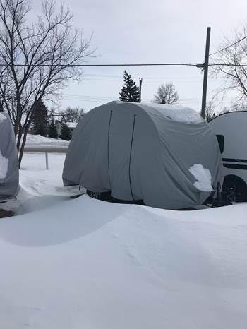 A small structure outdoors in a snowy area is protected by a gray OUT113 NuCamp TAG Trailer Cover from Paha Que Custom, surrounded by snowdrifts, bare trees, and a nearby RV under an overcast sky.