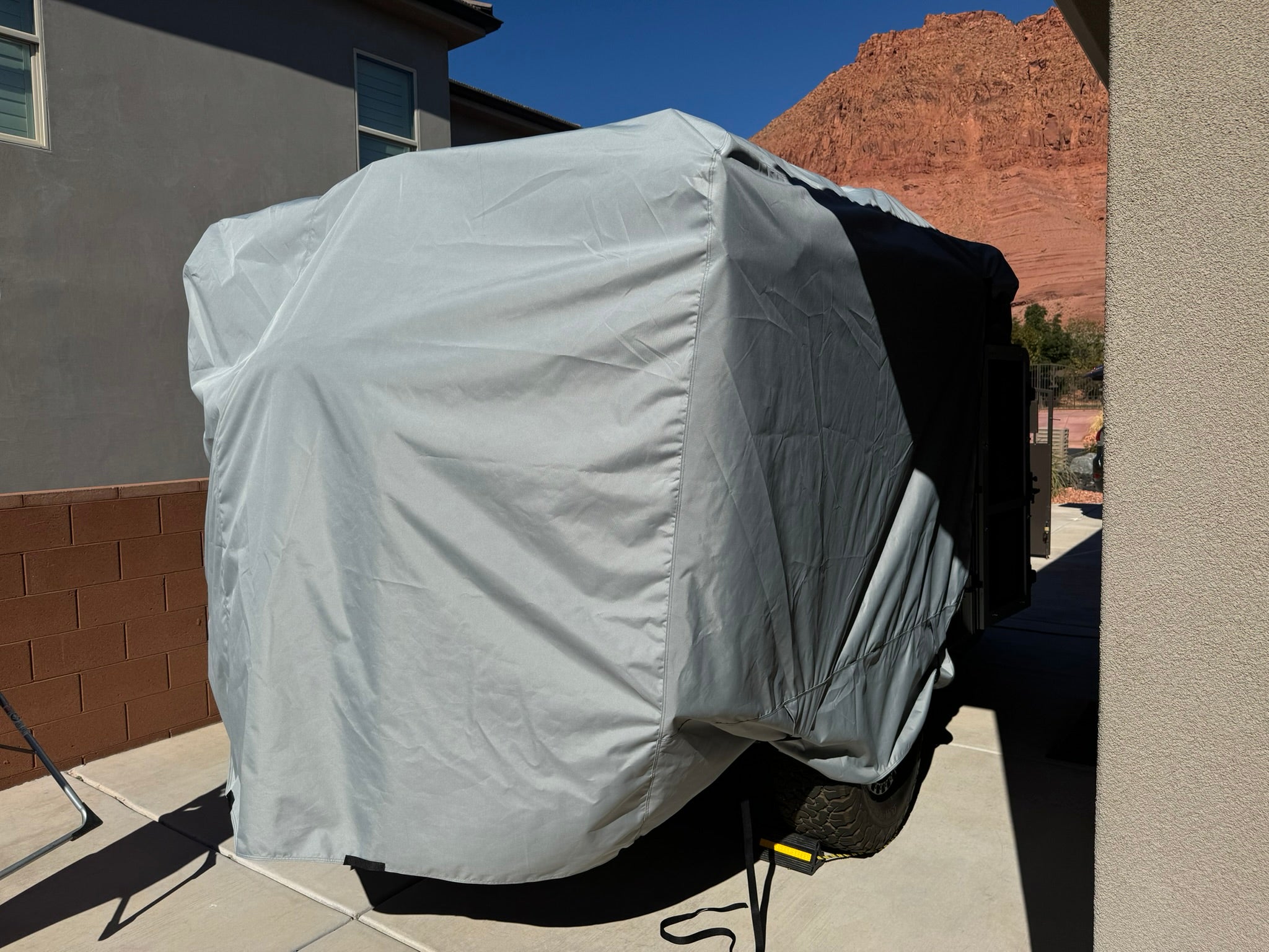 A large trailer is parked between two buildings, fully covered by the Paha Que Custom Teton X Trailer Cover Premium Protection for TetonX Trailers. Red rock hills and a clear blue sky are visible in the background.