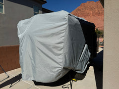A large trailer is parked between two buildings, fully covered by the Paha Que Custom Teton X Trailer Cover Premium Protection for TetonX Trailers. Red rock hills and a clear blue sky are visible in the background.