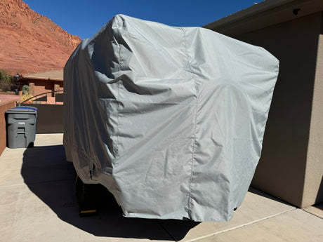 A large trailer is sheltered in a driveway by the Paha Que Custom Teton X Trailer Cover Premium Protection for TetonX Trailers, with a trash bin and red rocks visible in the background under a clear blue sky.