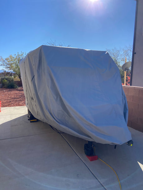 A large trailer is parked on a driveway, protected by the gray Paha Que Custom Teton X Trailer Cover Premium Protection for TetonX Trailers. The sunny yard has red gravel, bushes, and a brick wall.