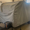 A UKAN Camp 590G/585G 5x9 Cover by PahaQue Wilderness, made of durable MagnaTuff fabric, protects a covered Little Guy Max trailer with a black storage box in a garage near a white vehicle, sunlight outside.
