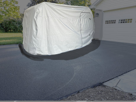 An Encore Trailer Cover by Paha Que Custom is suspended above a paved driveway in front of a white garage, framed by green trees and grass. This illustrates the premium protection offered by Encore Trailer Covers by PahaQue.