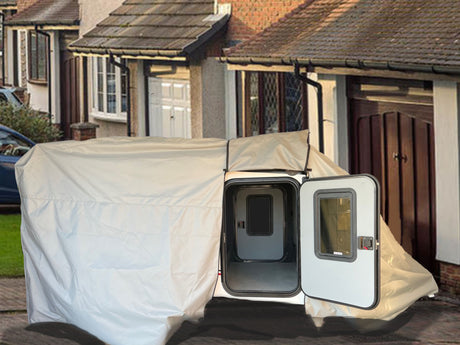 An Encore Trailer Cover by PahaQue Premium Protection from Paha Que Custom is set up on a driveway in front of suburban homes, protecting a small white trailer with an open door.