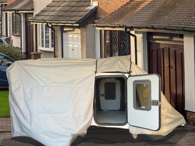 An Encore Trailer Cover by PahaQue Premium Protection from Paha Que Custom is set up on a driveway in front of suburban homes, protecting a small white trailer with an open door.