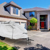 A trailer protected by the PahaQue Wilderness Zion Off Road Trailer Cover, made from heavy-duty MagnaTuff fabric, sits on the driveway before a large brick house with a red door, attached garage, and neat bushes.