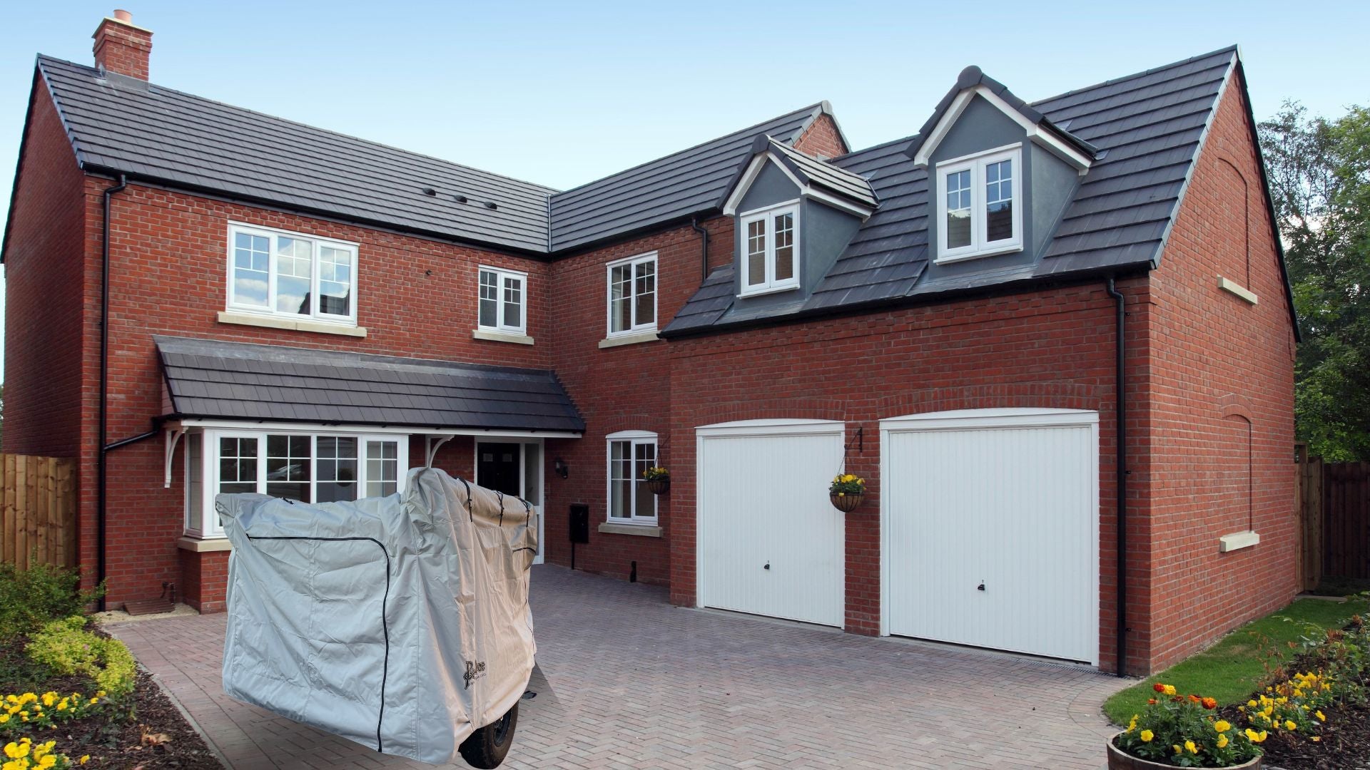 A modern two-story red brick house with white-framed windows, double garage doors, a covered motorcycle in the driveway, and a small flower garden. A PahaQue Wilderness Zion Off Road Trailer Cover (Heavy Duty MagnaTuff Fabric) provides protection from the elements.