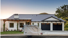 A modern house with a metal roof, white exterior, and two black garage doors has a covered trailer in the driveway featuring the Zion Off Road Trailer Cover Heavy Duty MagnaTuff Fabric by PahaQue Wilderness. Green grass, plants, a tree, and hills are in the background.