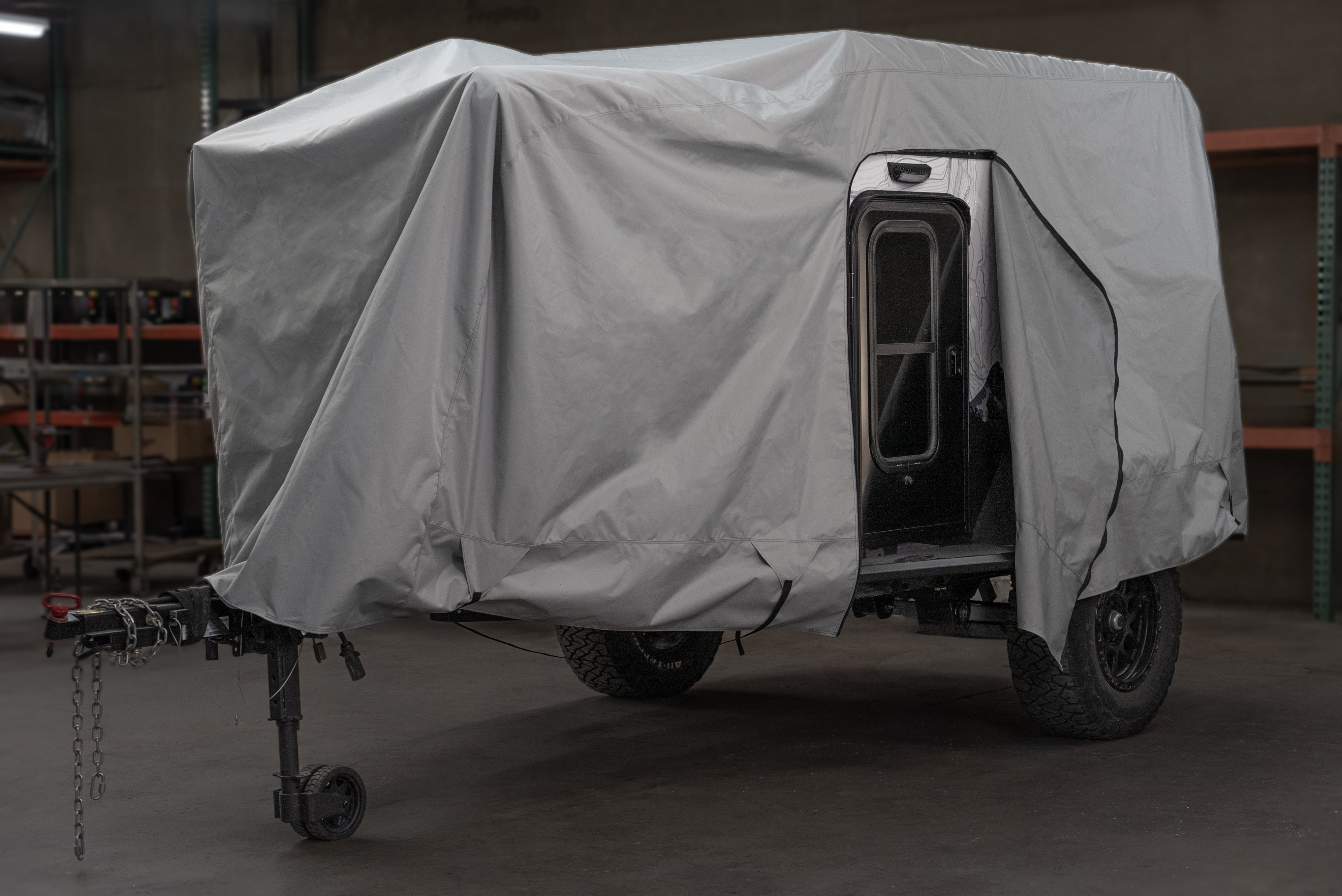 A black off-road camper trailer is partly covered with a gray Paha Que Custom Xpedition Trailer Cover for Ultimate Protection and Longevity inside a garage, with the side door open and shelving units visible in the background.
