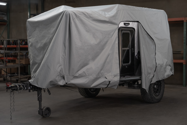 A black off-road camper trailer is partly covered with a gray Paha Que Custom Xpedition Trailer Cover for Ultimate Protection and Longevity inside a garage, with the side door open and shelving units visible in the background.