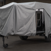 A black off-road camper trailer is partly covered with a gray Paha Que Custom Xpedition Trailer Cover for Ultimate Protection and Longevity inside a garage, with the side door open and shelving units visible in the background.