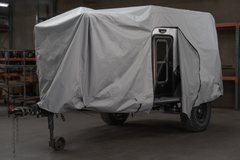 A black off-road camper trailer is partly covered with a gray Paha Que Custom Xpedition Trailer Cover for Ultimate Protection and Longevity inside a garage, with the side door open and shelving units visible in the background.