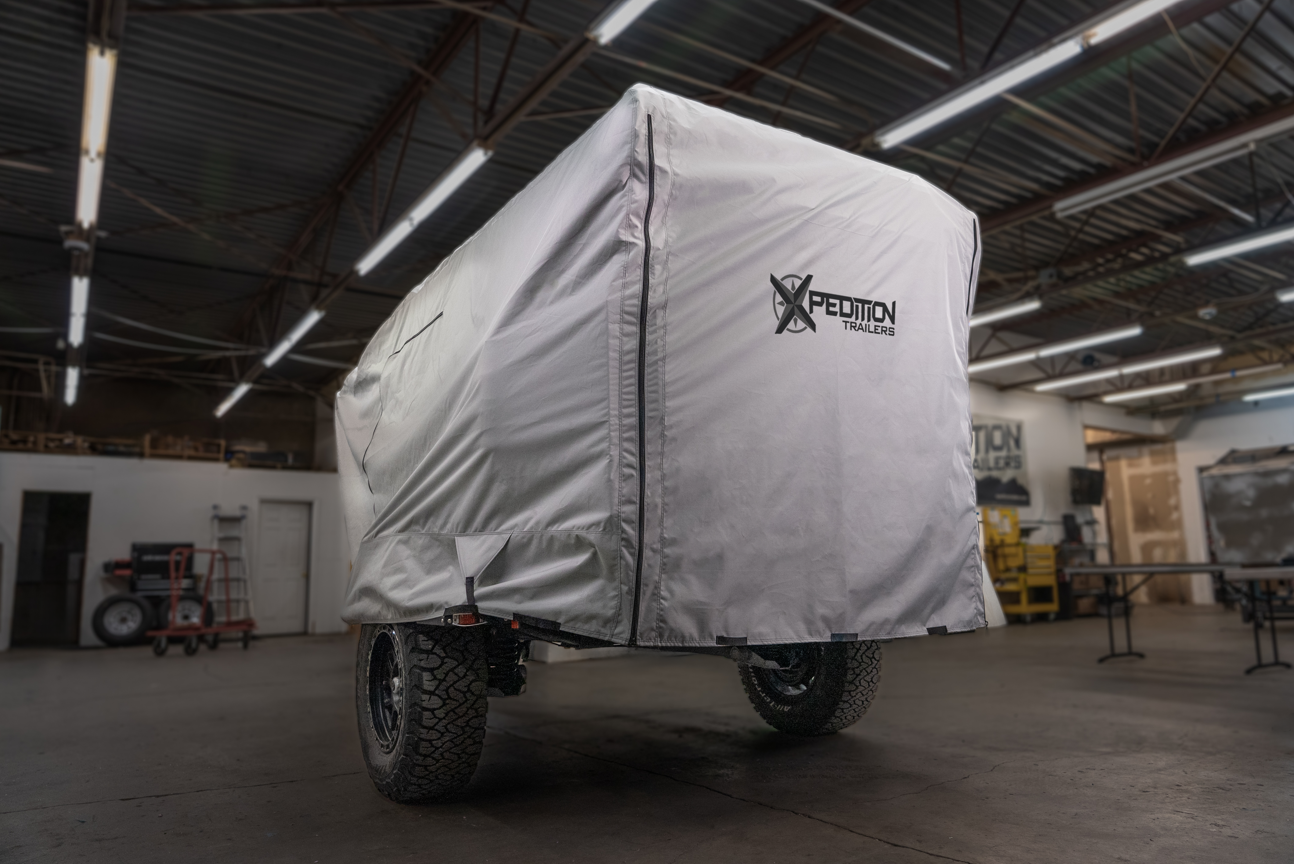 A covered off-road trailer with large tires is parked indoors under ceiling lights. The gray Xpedition Trailer Cover for Ultimate Protection and Longevity by Paha Que Custom displays the Expedition Trailers logo, with workshop tools and shelves in the background.