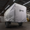 A covered off-road trailer with large tires is parked indoors under ceiling lights. The gray Xpedition Trailer Cover for Ultimate Protection and Longevity by Paha Que Custom displays the Expedition Trailers logo, with workshop tools and shelves in the background.
