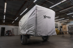 A covered off-road trailer with large tires is parked indoors under ceiling lights. The gray Xpedition Trailer Cover for Ultimate Protection and Longevity by Paha Que Custom displays the Expedition Trailers logo, with workshop tools and shelves in the background.