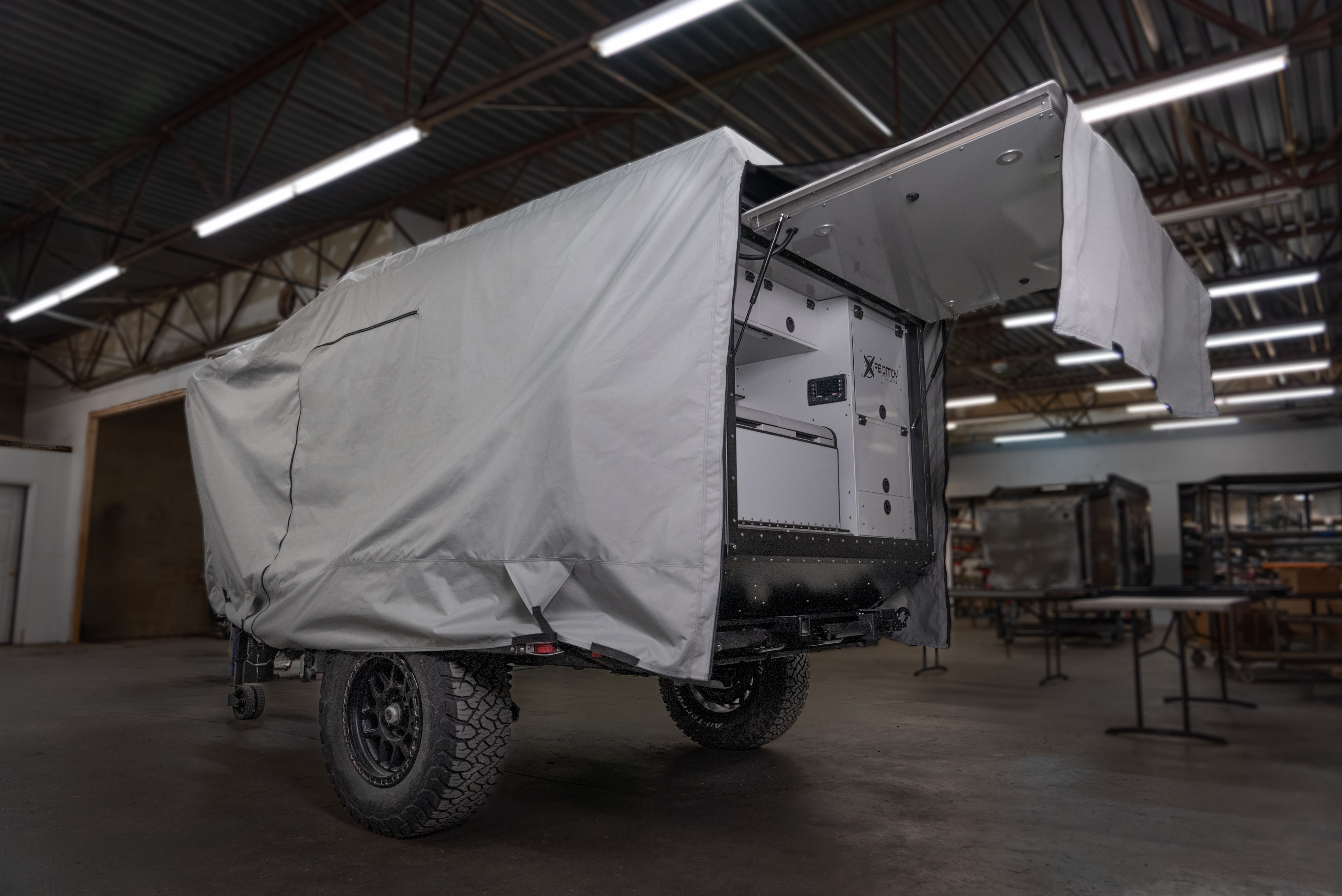 A rugged off-road trailer with large tires is partially covered by a Paha Que Custom Xpedition Trailer Cover for Ultimate Protection and Longevity, parked in a spacious industrial workshop. One end is open, showing storage compartments and protected interiors.