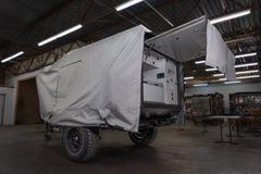 A rugged off-road trailer with large tires is partially covered by a Paha Que Custom Xpedition Trailer Cover for Ultimate Protection and Longevity, parked in a spacious industrial workshop. One end is open, showing storage compartments and protected interiors.