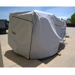 A Boreas Campers Trailer, protected by the Paha Que Custom Premium Trailer Cover, sits on a concrete driveway under clear skies, with trees and a fence in the background. The cover offers superior protection from the elements.