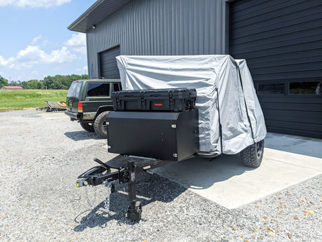 A black utility trailer with mounted storage boxes and a Paha Que Custom Cedar Ridge Vega XT AT Cover Premium Protective Solution for Cedar Ridge Campers is parked outside a gray metal building, near a dark SUV on a sunny gravel lot.