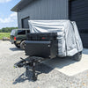 A black utility trailer with mounted storage boxes and a Paha Que Custom Cedar Ridge Vega XT AT Cover Premium Protective Solution for Cedar Ridge Campers is parked outside a gray metal building, near a dark SUV on a sunny gravel lot.