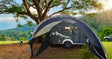 A gray camper with a mountain design sits in a grassy area beneath trees and mountains. A Paha Que Custom NuCamp TAB 320 Trailer Awning 2 by PahaQue—NEW FOR 2024—plus mesh side panels offers shade beside the trailer.