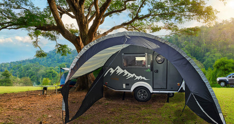 A gray camper with a mountain design sits in a grassy area beneath trees and mountains. A Paha Que Custom NuCamp TAB 320 Trailer Awning 2 by PahaQue—NEW FOR 2024—plus mesh side panels offers shade beside the trailer.