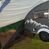 A gray teardrop camper with a mountain graphic is parked on grass, partly shaded by the NuCamp TAB 320 Trailer Awning 2 by Paha Que Custom—NEW FOR 2024. Green trees and blue sky are seen in the background.