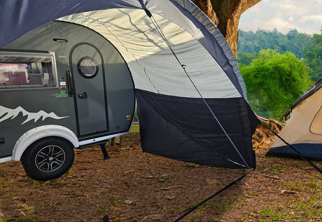 A small gray camper with a NuCamp TAB 320 Trailer Awning 2 by PahaQue, and a tan tent are set up under a large tree in a lush, green outdoor setting, partially framed by trees and grass. Brand: Paha Que Custom.