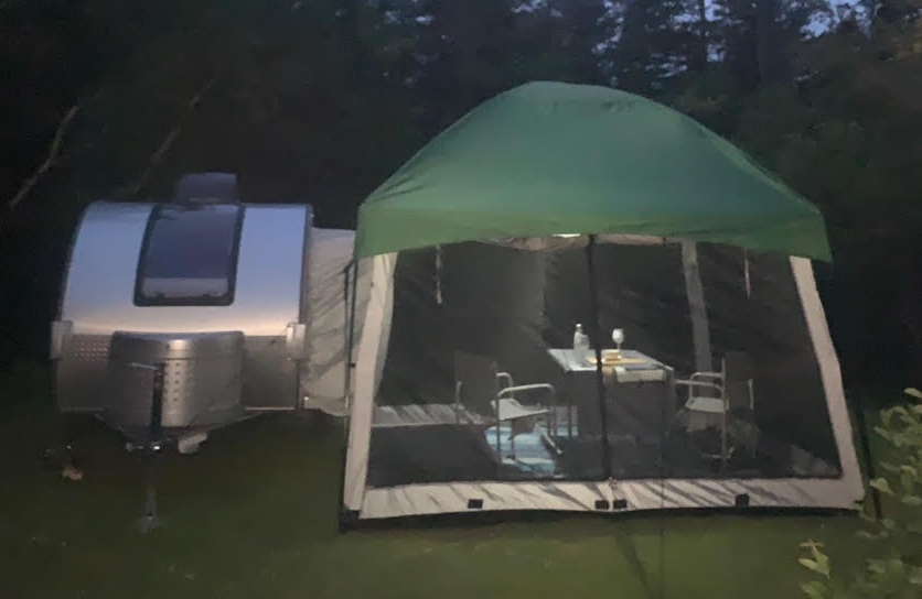 A small silver teardrop camper is parked beside a Paha Que Custom 5x7 Trailer Side Tent Screen Room for Camping and Outdoor Adventures, featuring two chairs and a table inside, surrounded by trees at dusk.