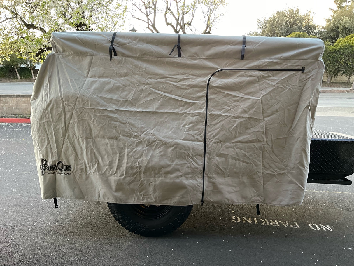 A tan PahaQue Wilderness Vintage Trailer Works XTR Cover, made from durable MagnaTuff fabric and featuring a zippered door, protects an outdoor grill or smoker mounted on a trailer with large black tires parked near a NO PARKING curb.