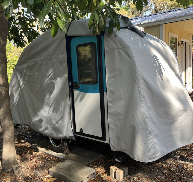 A Paha Que Custom Helio O Series Trailer sits under a tree beside a house, its blue trim and door visible beneath the Heavy Duty MagnaTuf Protection cover by PahaQue. Sunlight and shadows highlight the cover’s sturdy, weatherproof design.