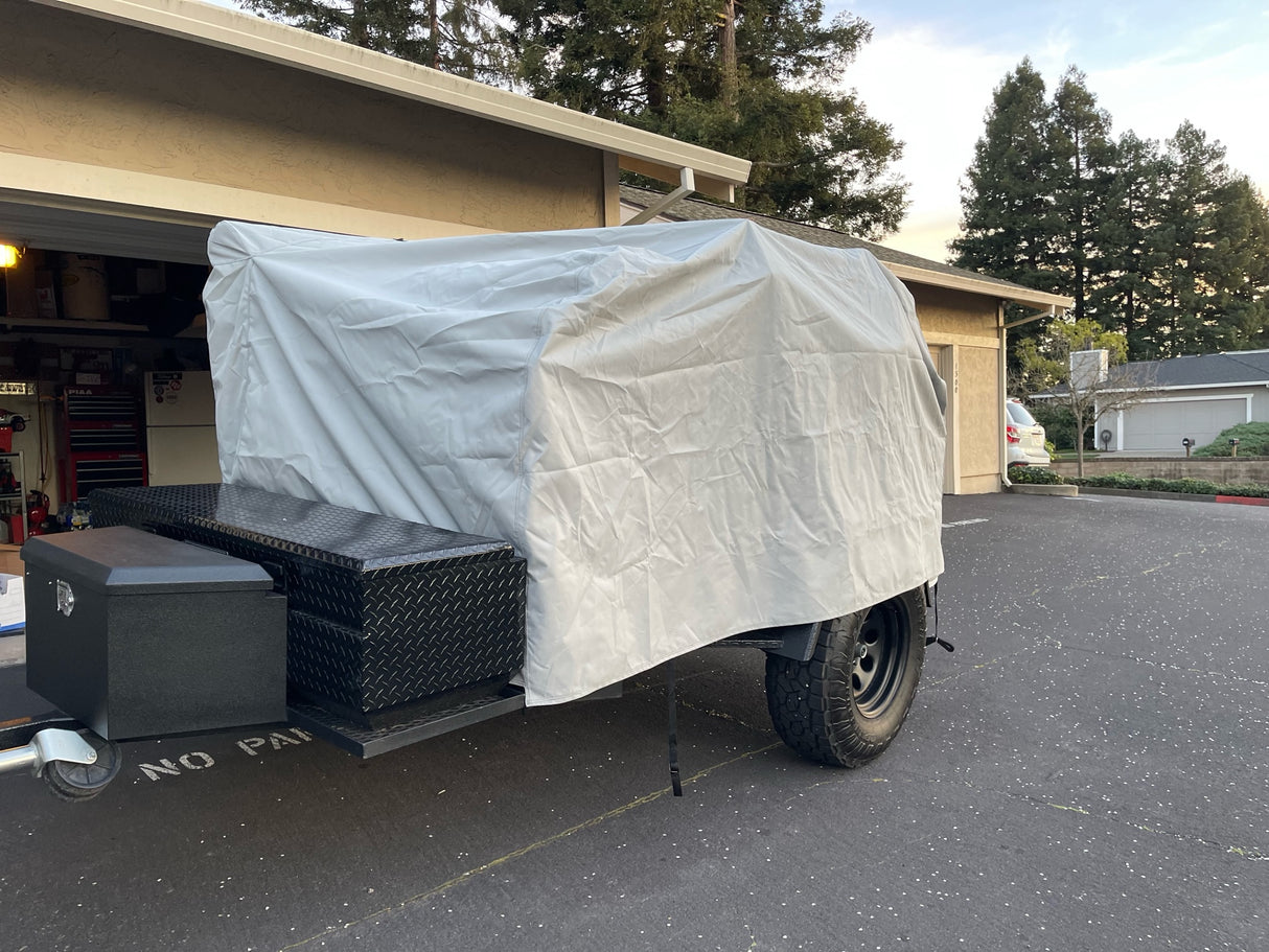 A small utility trailer is parked in a driveway, protected by the PahaQue Wilderness Vintage Trailer Works XTR Cover Heavy Duty Protection for Vintage Trailers. Trees and houses can be seen in the background on a clear day.