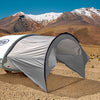 A small white teardrop camper with a Paha Que Custom NuCamp TAB 320 Trailer Awning is set up on a dry, rocky desert plain, its gray tent providing shade as tall mountains rise under a clear blue sky.