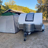 A gray teardrop camper with a Paha Que Custom 5x7 Trailer Side Tent Screen Room is parked by a large tent at a campsite surrounded by trees and a covered pavilion, with camping gear under the sunny, clear sky.