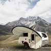 A compact white teardrop camper with a PahaQue Wilderness Little Guy Micro Max Visor awning is parked on grassland before rugged, snow-capped mountains under a partly cloudy sky.
